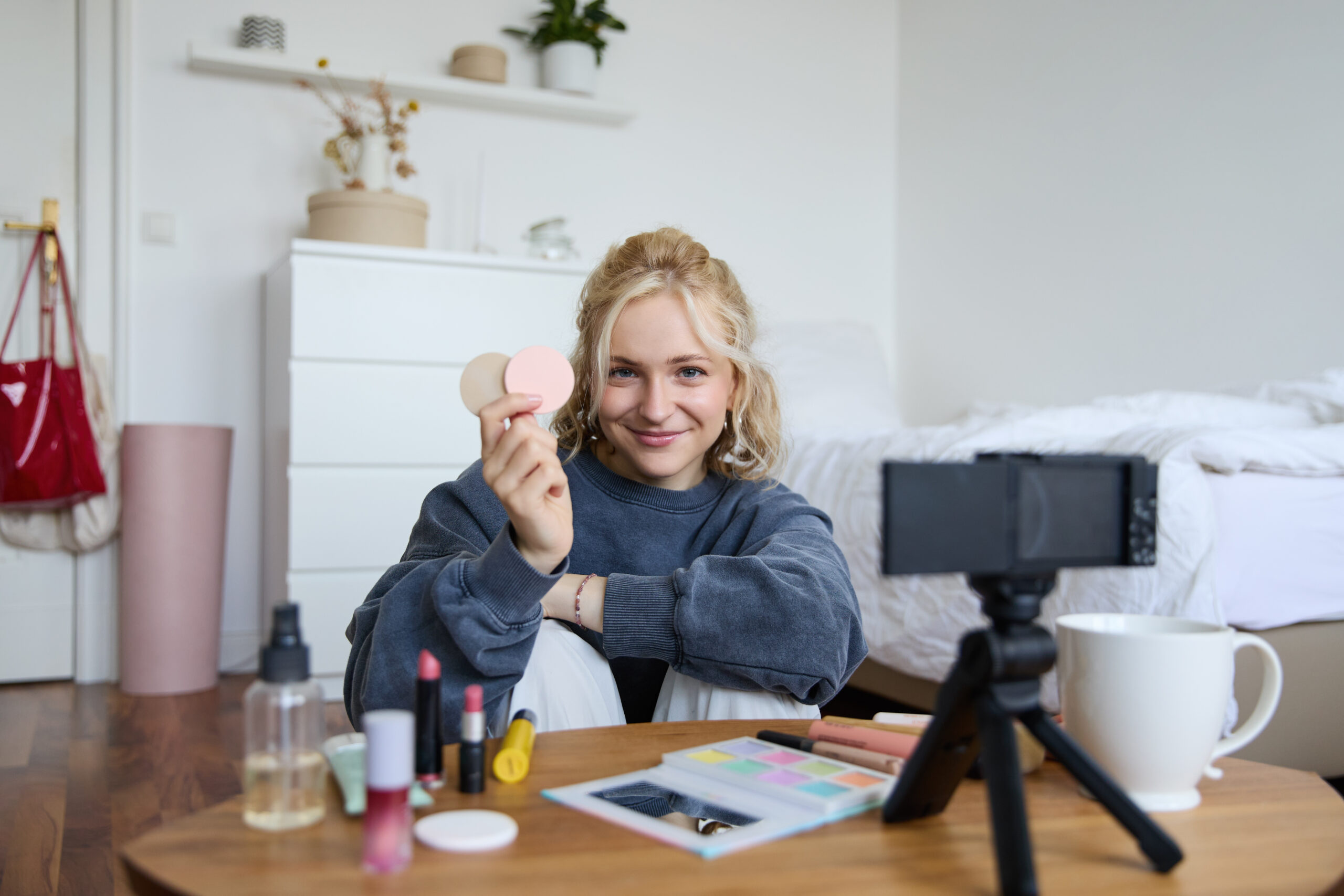 Eine junge Blonde Frau sitzt vor einem Smartphone und filmt sich und ihre Schmink Produkte